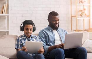 Gadget Generations. Afro father and son spending time with different electronic devices at home, man using laptop and boy holding digital tablet photo