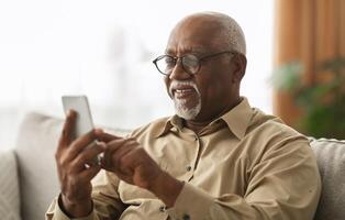 Senior Black Man Texting On Smartphone And Using Mobile Application Wearing Eyeglasses Sitting On Sofa At Home. Male Scrolling Reading News Feed In Social Media And Browsing Internet Via Phone photo