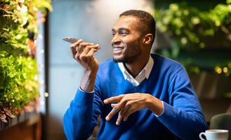 Voice Search. Happy black man using voice assistant on mobile phone or calling on smartphone at cafe, copyspace photo
