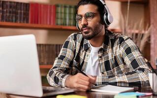 Arab Student Guy Learning Online At Laptop Computer Studying Sitting In Library Near Bookshelf In University. Modern Education, E-Learning And Online Training Concept. Selective Focus photo
