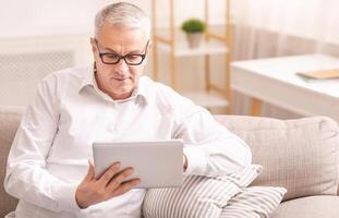 Reading News. Senior man in spectacles using digital tablet sitting on the couch at home, copyspace photo