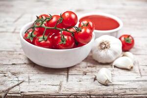 Fresh tomatoes in white bowl, sauce and raw garlic on rustic wooden table. photo