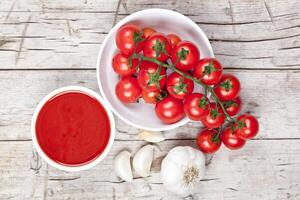 Fresh tomatoes in white bowl, sauce and raw garlic on rustic wooden table. photo