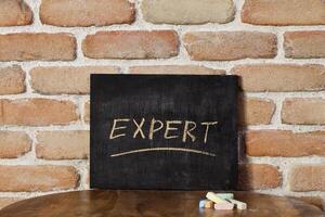 Chalk board with the word EXPERT drown by hand on wooden table on brick wall background. photo