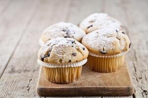 Four homemade fresh muffins with sugar powder and blueberries on cutting board on rustic wooden table. photo