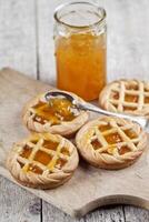 Fresh baked tarts with marmalade filling and apricot jam in glass jar on cutting board on rustic wooden table. photo