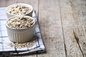 Oat flakes in ceramic bowls on linen napkin, golden wheat ears on rustic wooden background. photo
