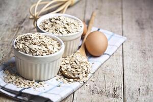 Oat flakes in ceramic bowls and wooden spoon and fresh chicken eggs on rustic wooden table background. photo