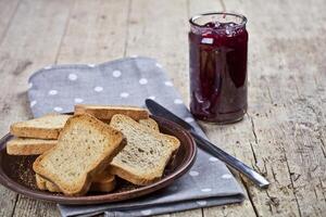 Toasted cereal bread slices on grey plate and jar with homemade cherry jam closeup on linen napkin on rustic wooden table background. photo