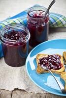Fresh cereal bread slices on blue ceramic plate, homemade cherry and wild berries jam in jars and spoons closeup on linen napkin on rustic wooden table background. photo