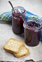 Toasted cereal bread slices on blue ceramic plate and homemade cherry and wild berries jam in jars and spoons closeup and linen napkin on rustic wooden table background. photo