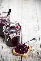 Fresh cereal bread slices and jars with homemade wild berries and cherry jam and spoons closeup on rustic wooden table background. photo