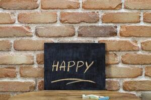 Chalk board with the word HAPPY drown by hand and chalks on wooden table on brick wall background. photo