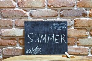 Chalk board with the word SUMMER drown by hand and chalks on wooden table on brick wall background. photo