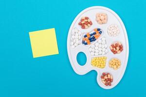 Global Pharmaceutical Industry and Medicinal Products - Different Colored Pills, Tablets and Capsules on White Art Palette with Blank Sticky Note and Copy Space Lying on Blue Background, Flat Lay photo