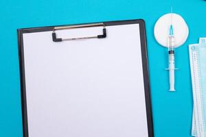 Vaccination, Immunology or Revaccination Concept - A Medical Syringe Lying on Blue Table in Doctor's Office in a Hospital or Clinic. Black Clipboard with Sheet of Paper - Mock Up with Copy Space photo