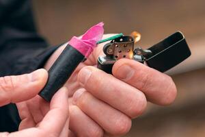 Setting Fire to the Firecracker. Man in Black Clothes Lighting Up the Petard. Firing Up the Pyrotechnics with a Black Gasoline Lighter Outdoors photo