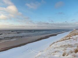 The beach is covered in snow except for some areas near the water, and there are small waves in the sea under a partly cloudy sky, which makes it an interesting contrast between winter and coastal photo