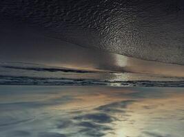 The image shows an inverted coastal scene during autumn, with the sky reflected on the water is surface, creating a unique visual effect. The tranquil beach and cloudy sky add to the serene atmosphere photo
