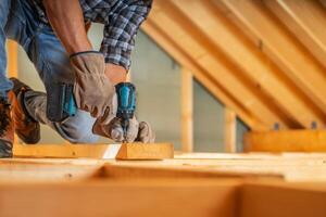 A Carpenter Uses a Power Drill to Secure Beams in a Daylight Attic Renovation photo