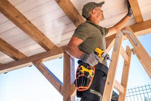 Skilled Worker Installing Ceiling Paneling While Standing on Ladder in a Sunny Outdoor Workspace photo