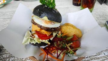 Gourmet black charcoal burger with egg, cheese, and vegetables, served with crispy wedges and fresh salad on parchment, creating a vibrant and appetizing presentation. photo