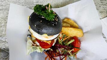 Gourmet black charcoal burger with egg, cheese, and vegetables, served with crispy wedges and fresh salad on parchment, creating a vibrant and appetizing presentation. photo