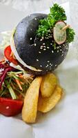 Gourmet black charcoal burger with egg, cheese, and vegetables, served with crispy wedges and fresh salad on parchment, creating a vibrant and appetizing presentation. photo