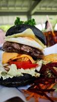 Gourmet black charcoal burger with egg, cheese, and vegetables, served with crispy wedges and fresh salad on parchment, creating a vibrant and appetizing presentation. photo