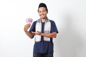 Portrait of frustrated Asian muslim man wearing sarung scarf showing one hundred thousand rupiah while pointing to the side. Financial and investment concept. Isolated image on white background photo