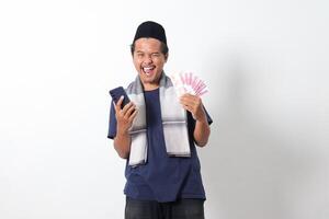 Portrait of excited Asian muslim man wearing sarung scarf holding rupiah money banknote while holding and looking at his mobile phone. Isolated image on white background photo
