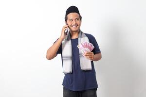 Portrait of excited Asian muslim man wearing sarung scarf holding one hundred thousand rupiah while having a conversation on his mobile phone. Isolated image on white background photo