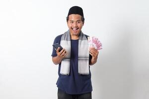 Portrait of excited Asian muslim man wearing sarung scarf holding rupiah money banknote while holding and looking at his mobile phone. Isolated image on white background photo