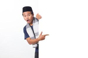 Portrait of excited Asian muslim man wearing sarung scarf hiding behind empty white billboard with mockup template and copy space for graphic. Advertising poster. Isolated on white background photo