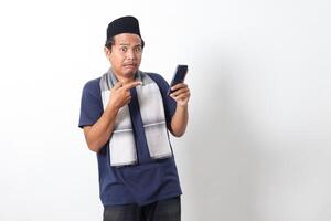 Portrait of upset Asian muslim man wearing sarung scarf and skullcap, holding mobile phone and showing shocked expression. Isolated on white background photo