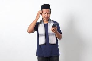 Portrait of shocked Asian muslim man wearing sarung scarf and skullcap, holding mobile phone and showing disappointed expression. Isolated on white background photo