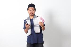 Portrait of shocked Asian muslim man wearing sarung scarf holding rupiah money banknote while holding and looking at his mobile phone. Isolated image on white background photo