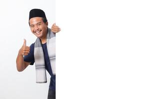 Portrait of excited Asian muslim man wearing sarung scarf hiding behind empty white billboard with mockup template and copy space for graphic. Advertising poster. Isolated on white background photo