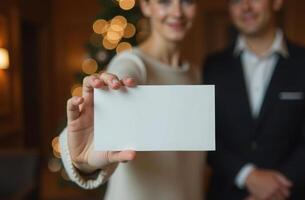 A woman holds a blank card in the foreground. in the background, a softly focused man and a christmas tree with twinkling lights create a warm, festive ambiance. photo
