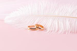 Pair of gold wedding rings on a pink background with a white decorative light feather. A copy space. The concept of the wedding. layout for design. photo