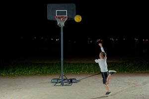 A child shoots a basketball at a hoop during nighttime practice in an outdoor playground with city lights in the background photo
