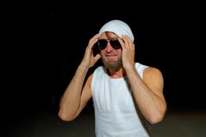 A man wearing a tank top, sunglasses, and a beanie poses confidently at night against a dark background photo