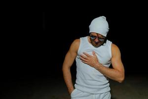 A man wearing a tank top, sunglasses, and a beanie poses confidently at night against a dark background photo