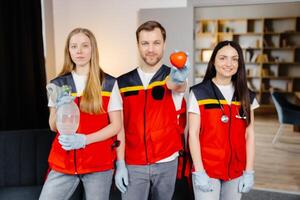 A group of young medical people in uniform stand in the middle of the classroom and smile after a first aid lesson. A man holds a red heart in his hands. Concept of assistance photo