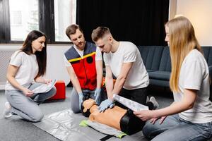 Young man instructor helping to make first aid heart compressions with dummy during the group training indoors photo