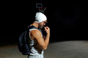 A muscular man wearing sunglasses and a beanie while holding backpack stands confidently in the dark photo