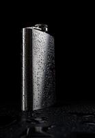 Stainless steel flask on a black background with water drops.Personal flat flask. photo