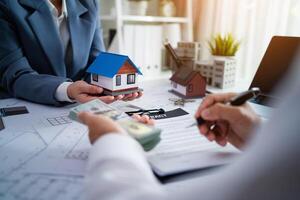 A man and a woman are sitting at a table with a house model and a stack of cash. The woman is holding the house model and the man is writing something on a piece of paper photo