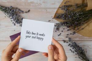 Female hands taking paper card note with text TAKE TIME TO MAKE YOUR SOUL HAPPY from violet envelope. Lavender flower. Top view, flat lay. Concept of mental spiritual health self care wellbeing mindfulness photo
