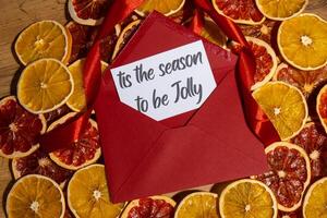 Christmas handmade wreath made of dried slices of orange and grapefruit with red envelope and paper note with IT'S THE SEASON TO BE JOLLY text. Winter holiday celebration greeting card. top view photo